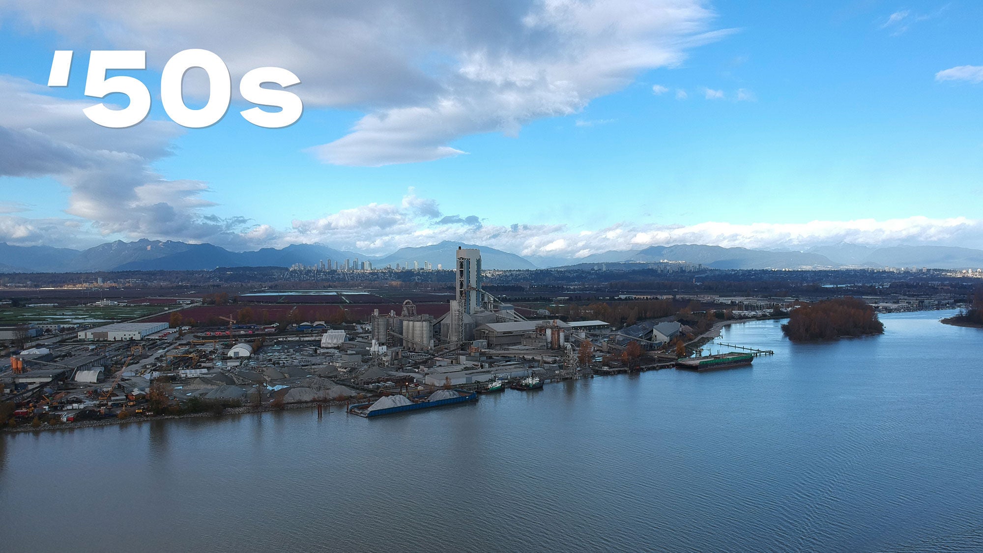 Aerial view of an industrial complex and cement plant by a wide river under a cloudy blue sky. Mountains and the city skyline are visible in the distance. There is "’50s" in bold white text above.