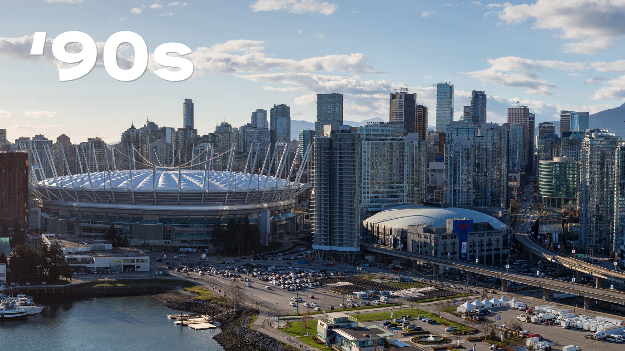 Aerial view of a cityscape with two large stadiums and modern skyscrapers under a blue sky with scattered clouds. A waterfront and roadways are visible in the foreground. There is “2000s” in bold white text above.