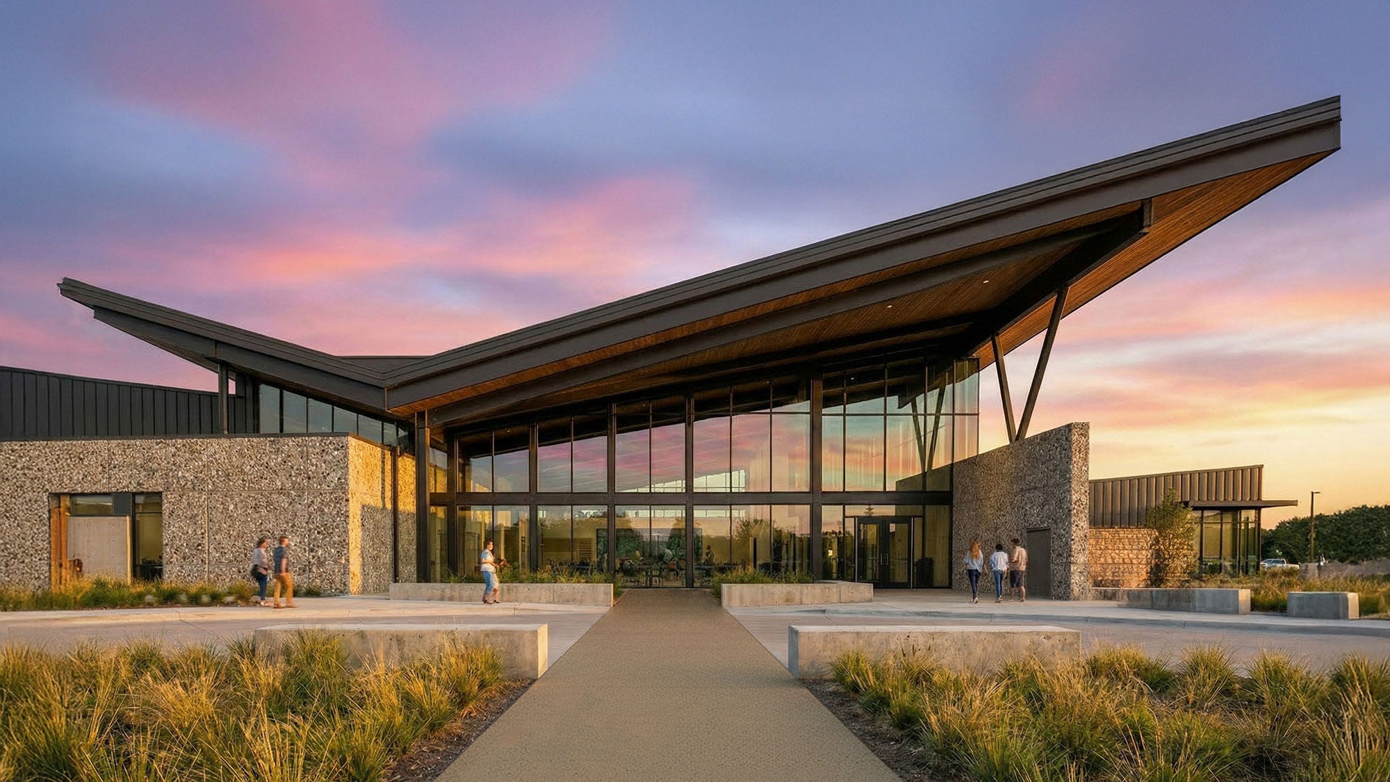 A glass and concrete recreational center with a large angled roof seen at dusk