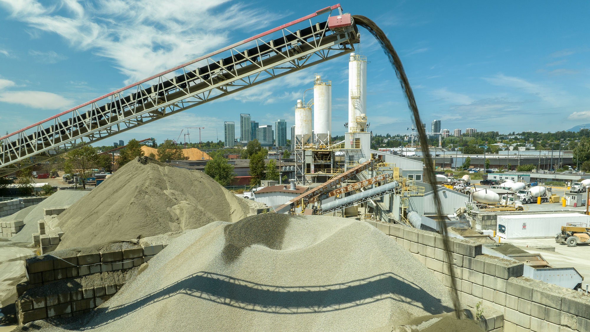 Aerial view of a concrete plant by a river, featuring storage silos, large piles of gravel, concrete trucks, and industrial buildings, with lush greenery and bridges in the background.