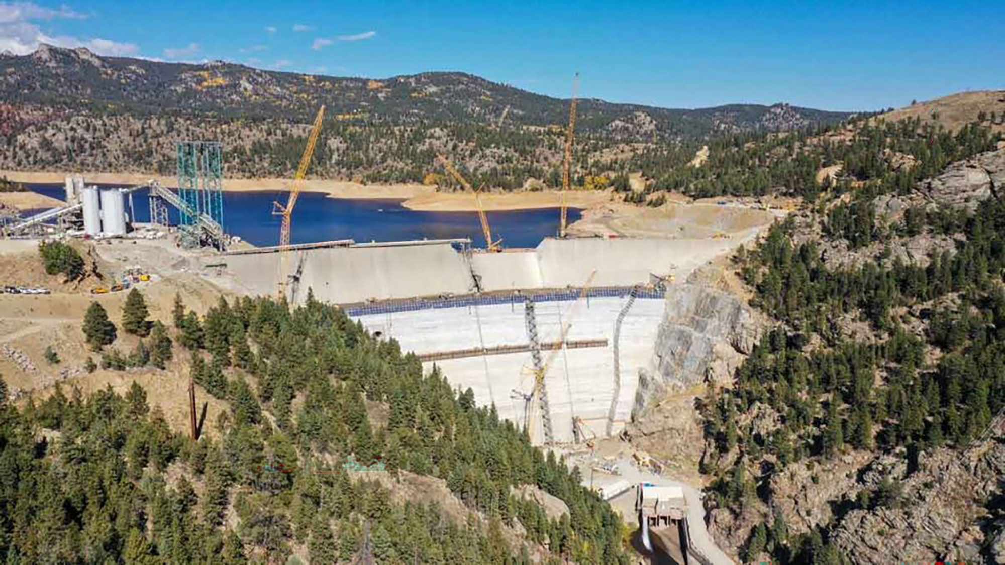 Vue aérienne d’un barrage en construction dans une zone montagneuse, entourée de collines forestières. Les grues sont visibles et un réservoir se forme derrière le barrage.
