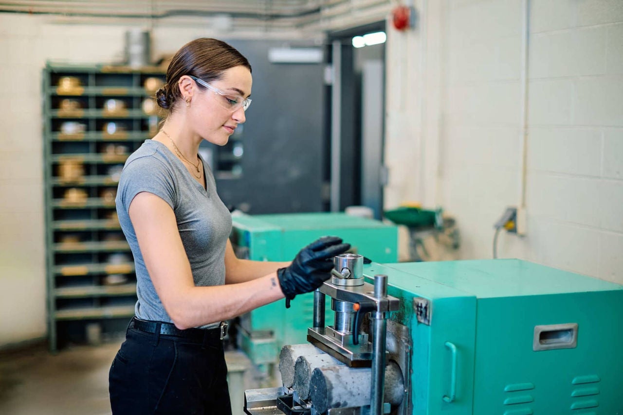 Une jeune femme utilisant une machine dans un atelier, portant des gants et des lunettes de protection
