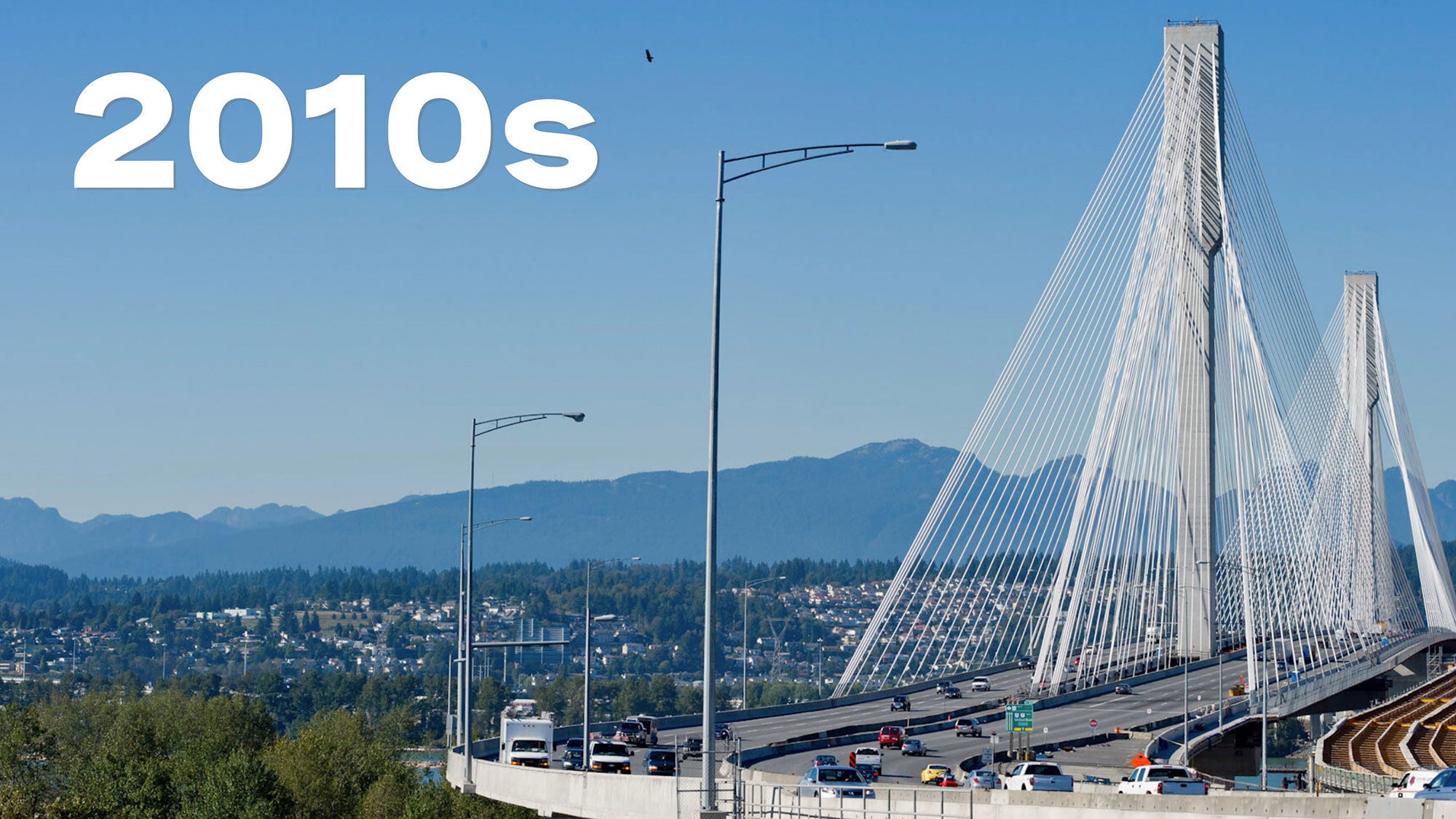 A suspension bridge spans across a river with vehicles traveling on it. The background features distant mountains against a clear blue sky. The word “2010s” is in bold white text above.