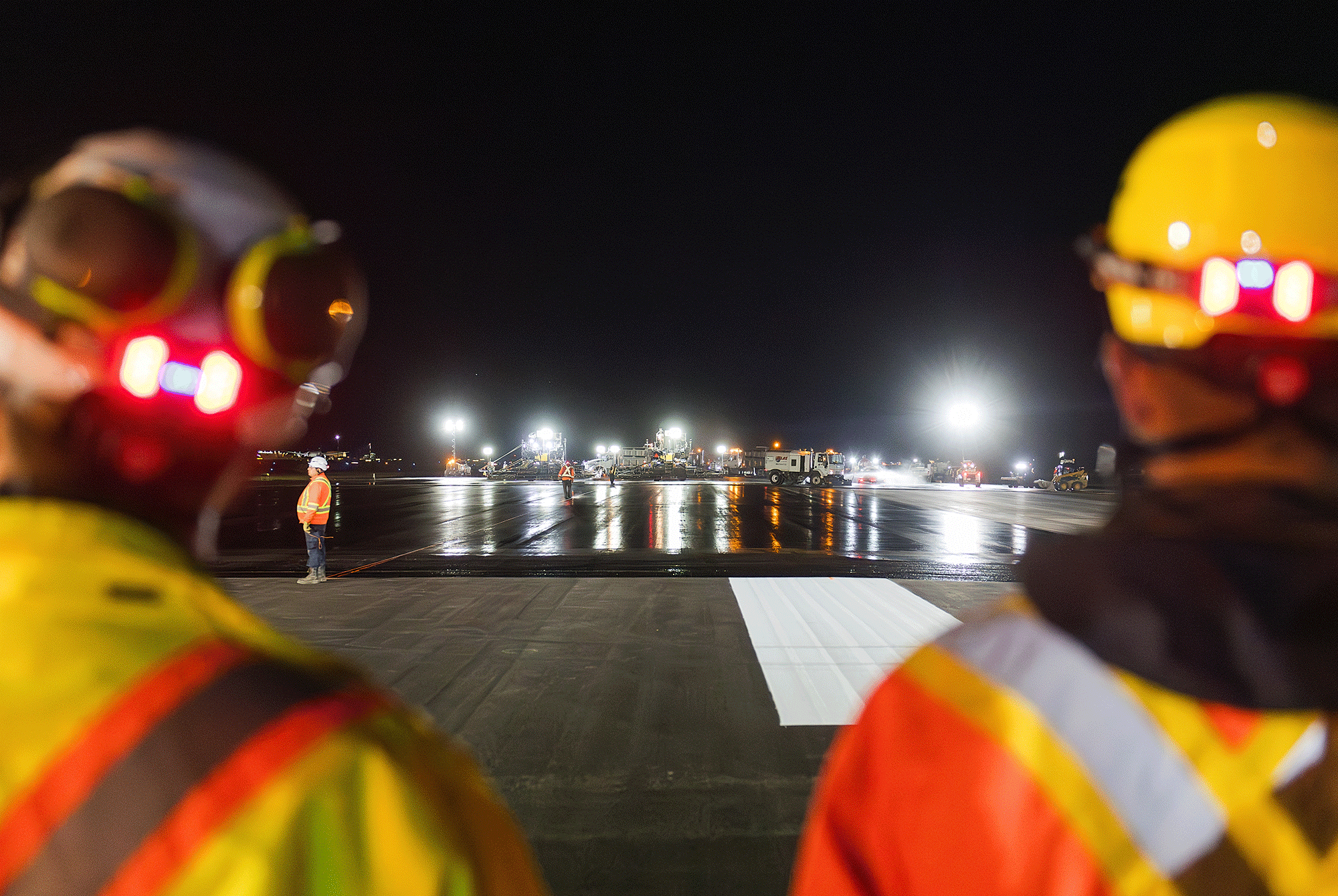 Two construction workers in safety gear and hard hats observe a brightly lit tarmac at night, creating an atmosphere of focus and diligence.
