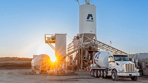 A large materials silo with an Amrize branded mixing truck beneath it at sunrise