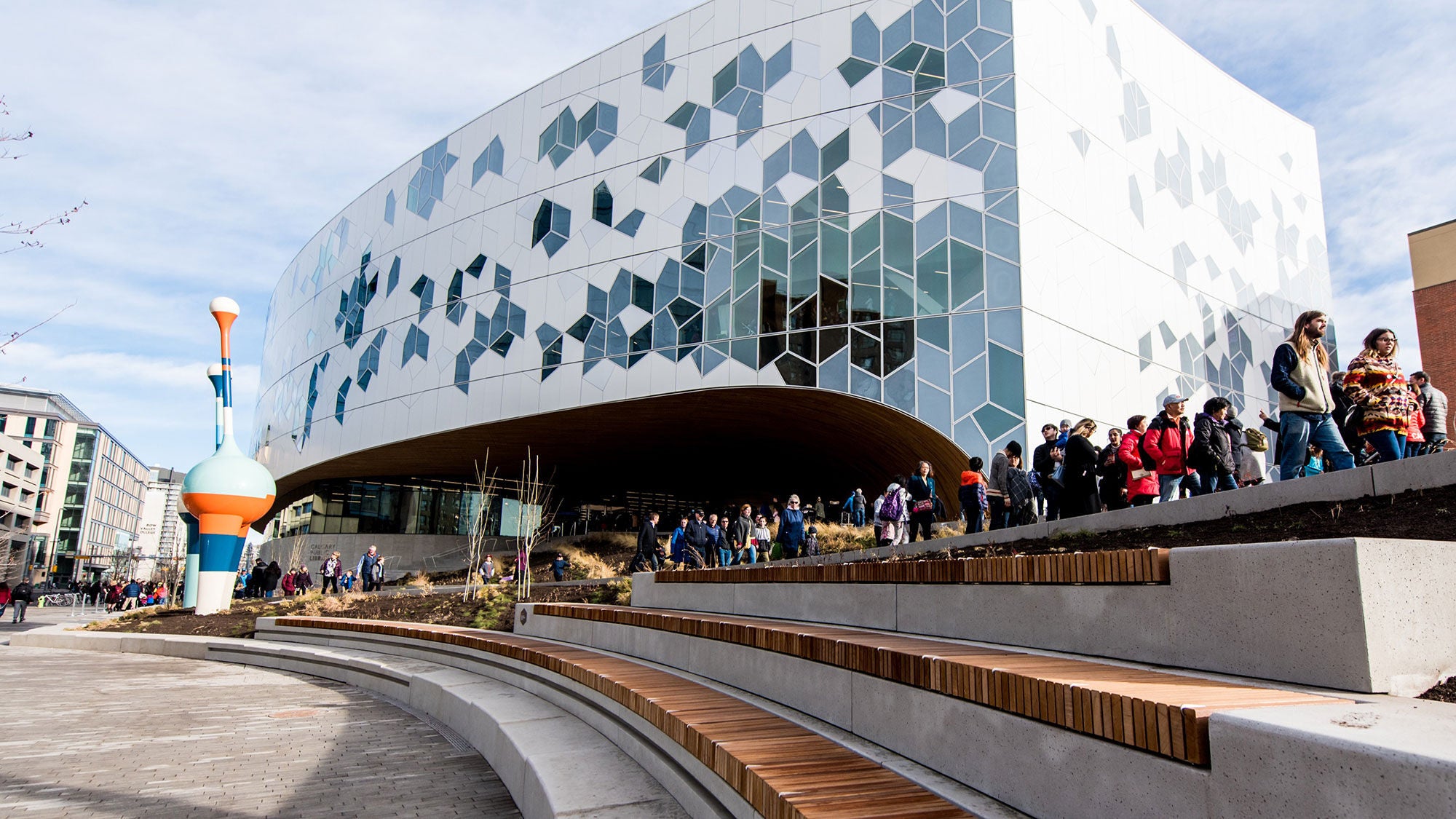 Low-angle view of the Calgary Central Library's outdoor plaza, highlighting tiered concrete and benches with crowds of people walking near the building.