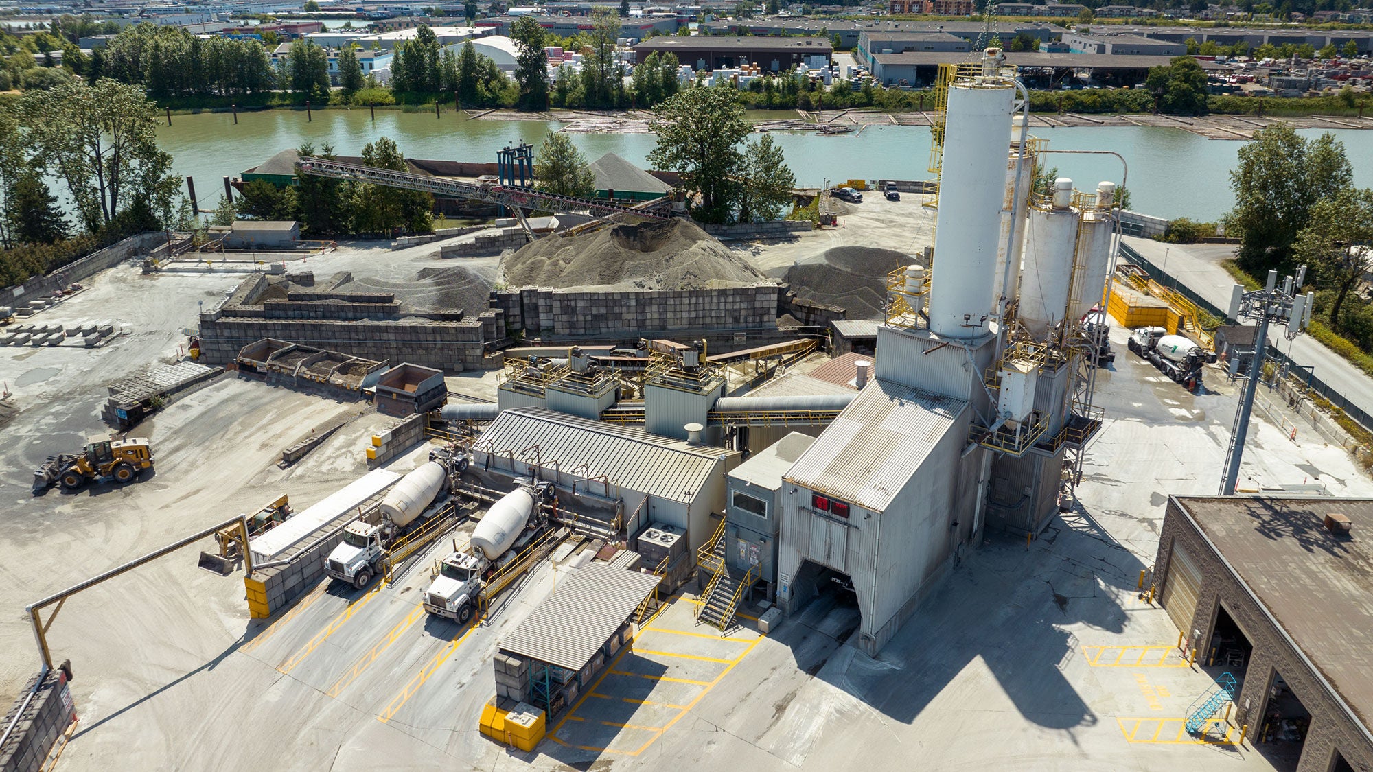 Aerial view of a concrete plant by a river, featuring storage silos, large piles of gravel, concrete trucks, and industrial buildings, with lush greenery and bridges in the background.