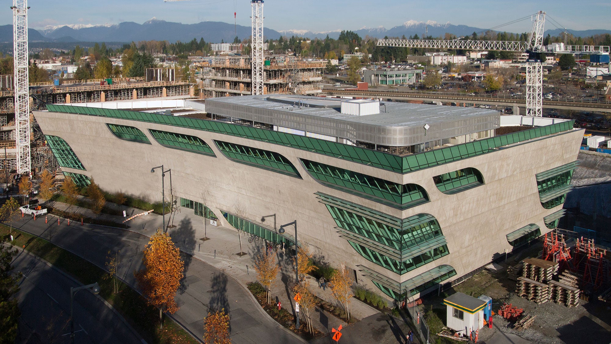 A photo of a multi-story modern library under construction and surrounded by cranes with snow-capped mountains in the distant background.