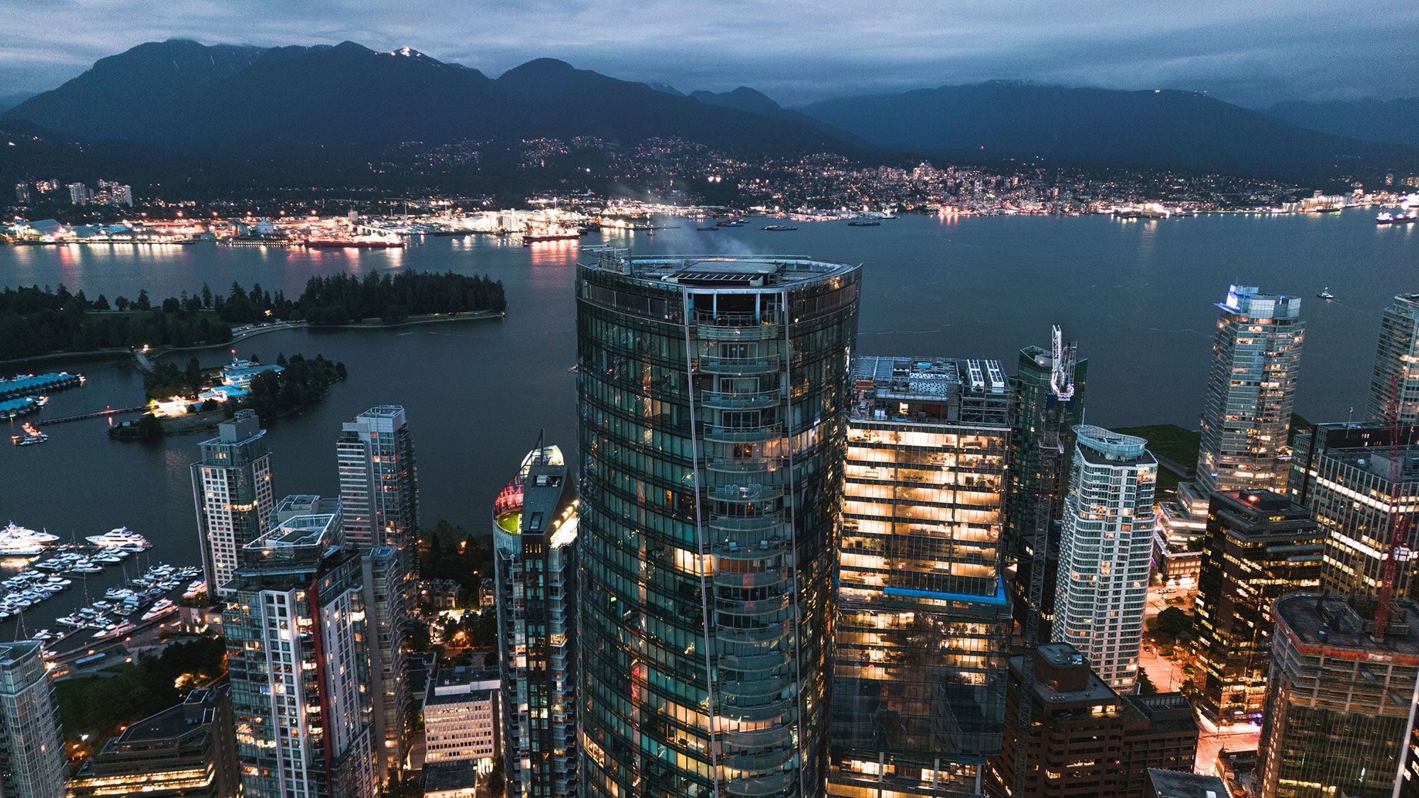 Aerial view of a cityscape at dusk, featuring illuminated skyscrapers near a body of water, with dark mountains and scattered city lights in the background.