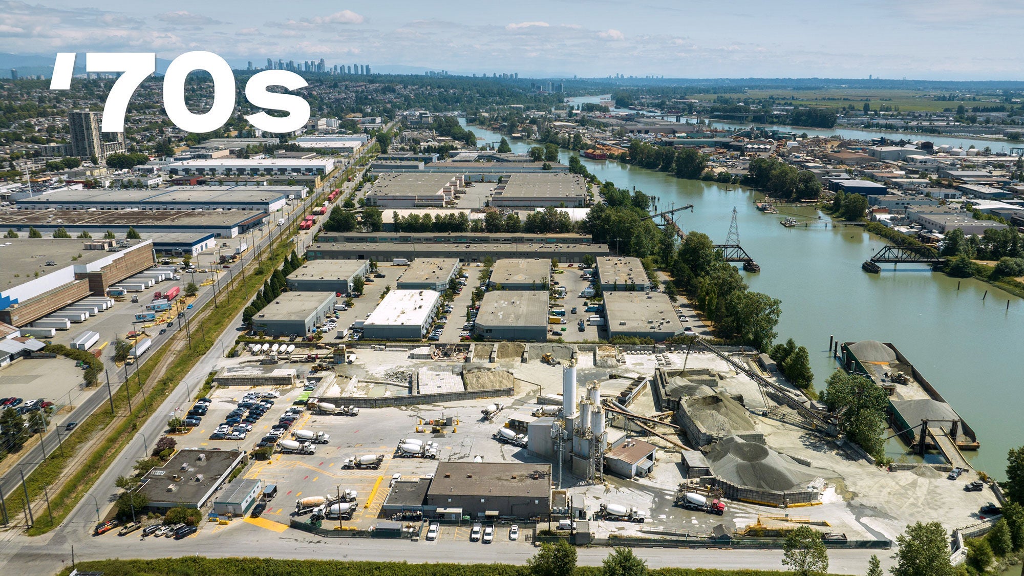 Aerial view of an industrial area with several large warehouses and a river. The skyline and distant cityscape are visible under a clear blue sky. There is "’70s" in bold white text above.