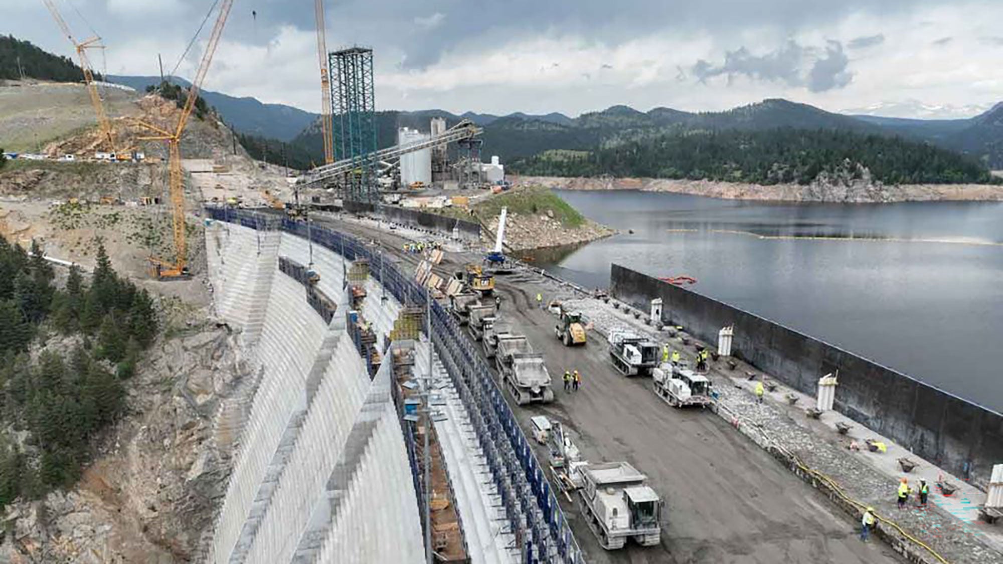 Vue aérienne d’un chantier de construction de barrage avec des grues et de la machinerie par une journée nuageuse. Le barrage partiellement construit suit une courbe le long d’une colline rocheuse près d’un grand réservoir.