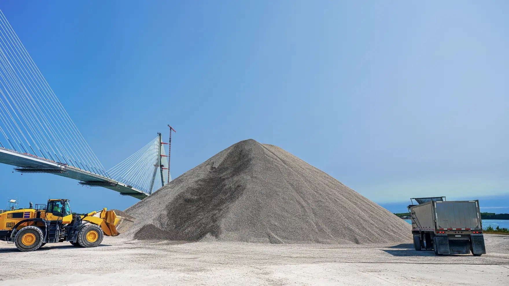 Pile of aggregate, construction trucks, Gordie Howe Bridge in the background 