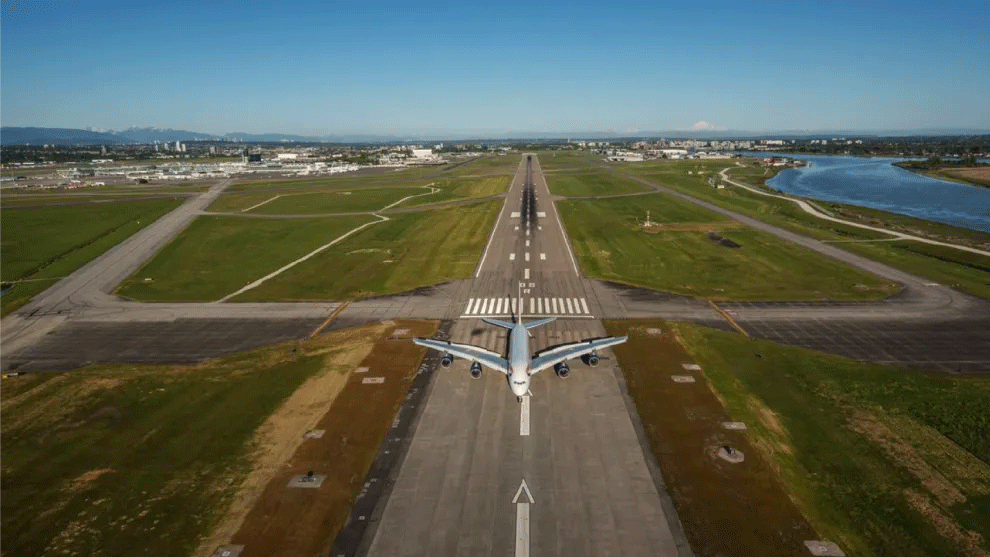 A plane sits on a runway with a river to the side and a city behind